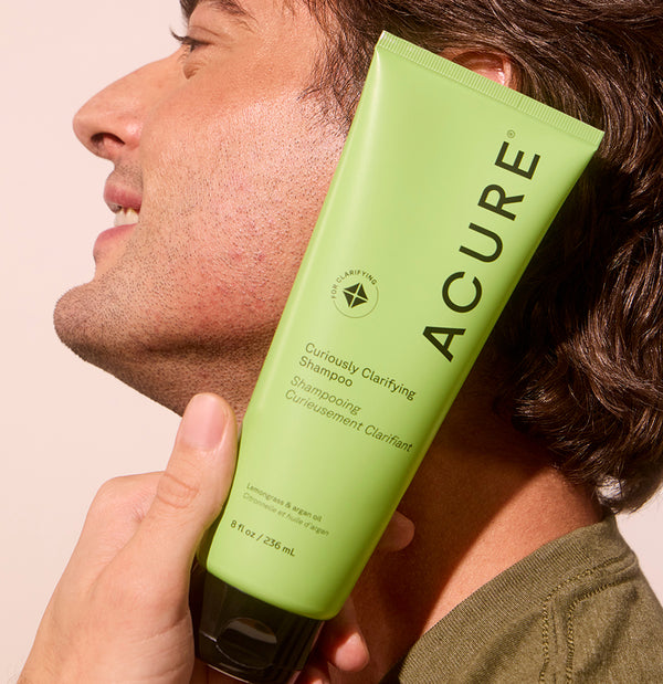Person holding a green Acure shampoo bottle near their head against a beige background